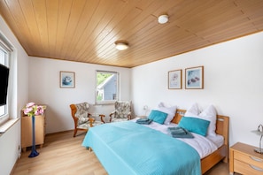 A bedroom with a wooden ceiling, a bed with blue bedding, a chair, a small table with a vase of flowers, and framed pictures on the wall.