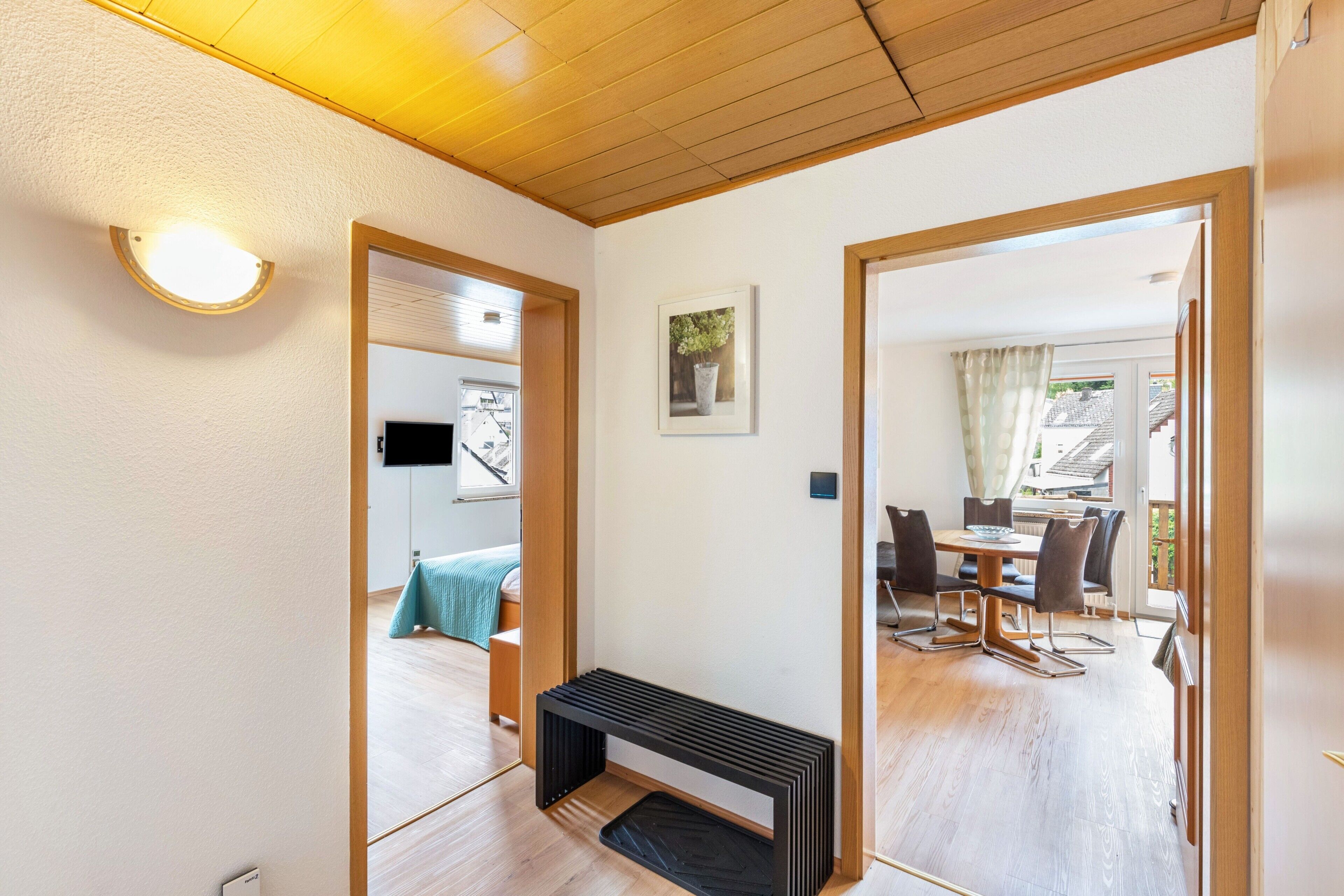 Un pasillo con techo de madera, un banco, un cuadro enmarcado y una zona de comedor con sillas y una mesa.