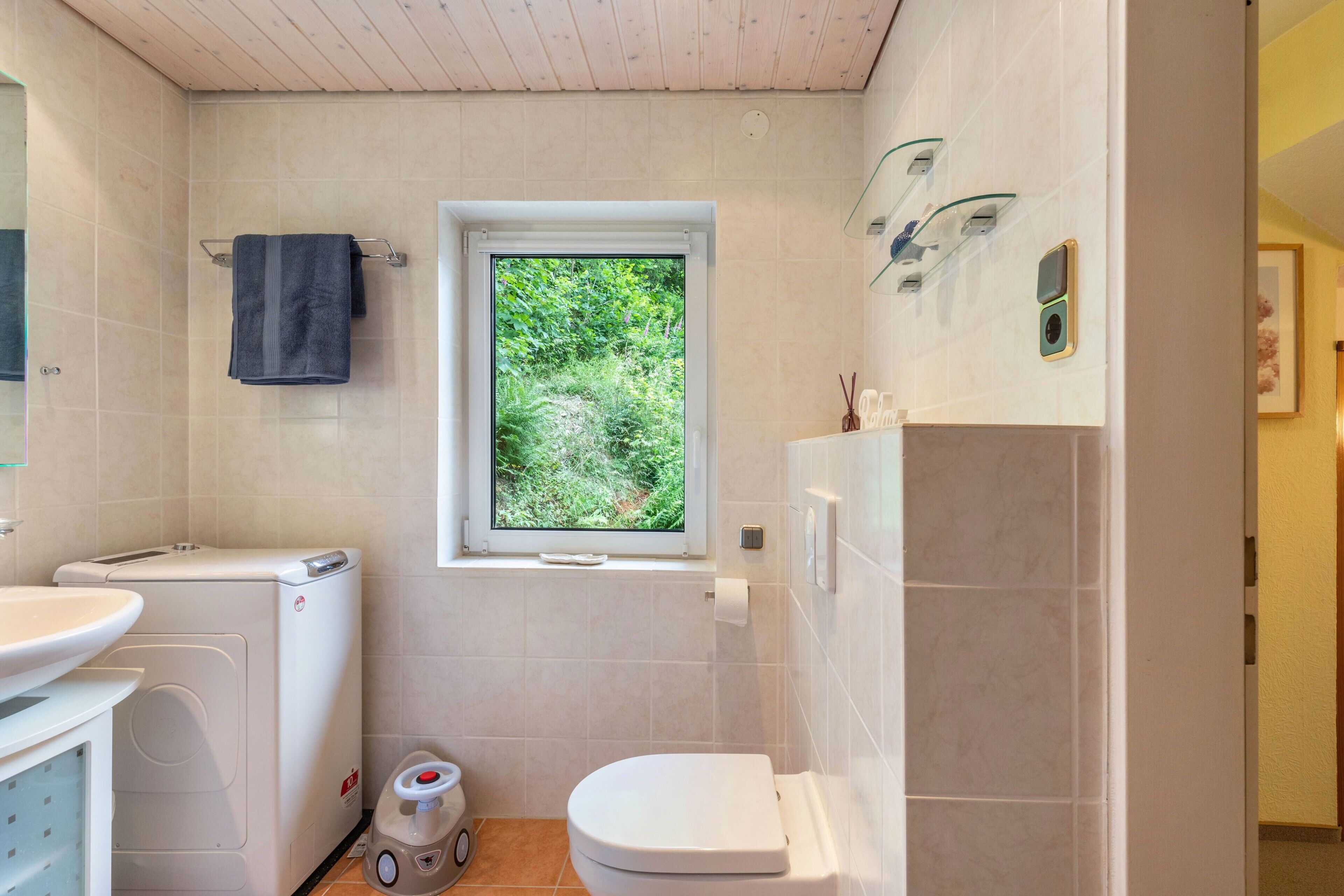 Une salle de bain comprenant des toilettes, un lave-linge, un lavabo, une fenêtre donnant sur un espace verdoyant et un espace douche.
