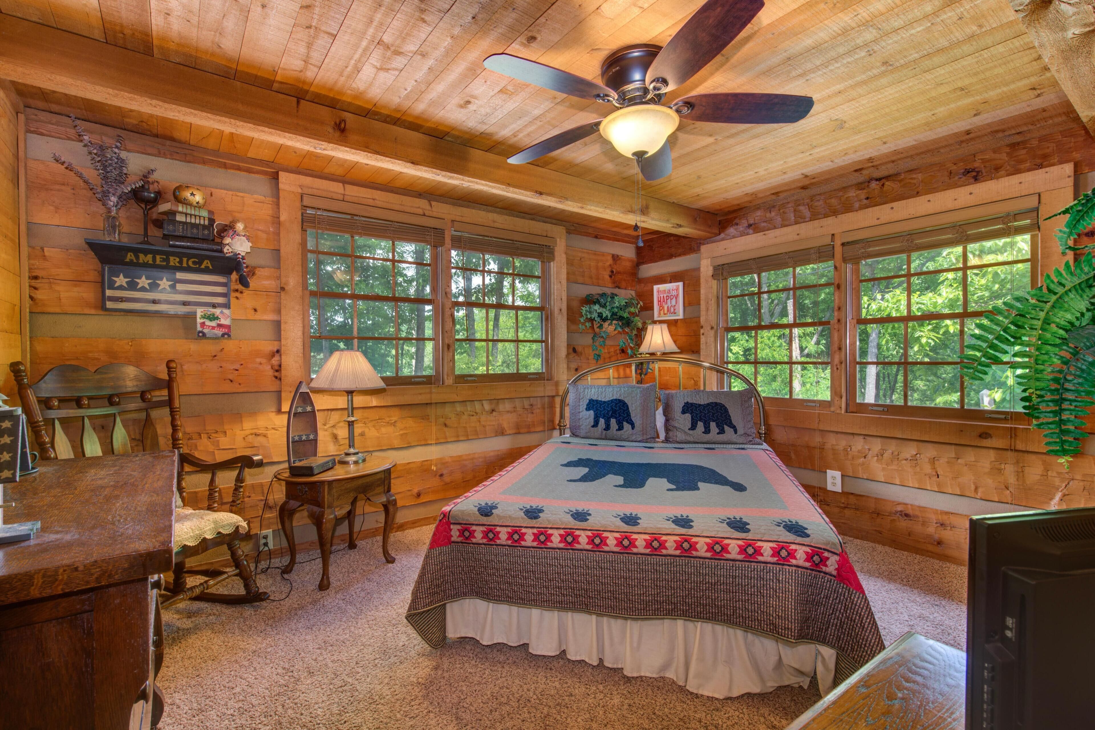 Dormitorio de cabaña de madera con cama, ventilador de techo, escritorio y ventana con vista a árboles.