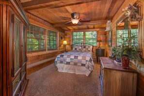 Ein Schlafzimmer in einer Holzhütte mit Bett, Nachttisch, Kommode, Deckenventilator und Fenster mit Fensterläden.