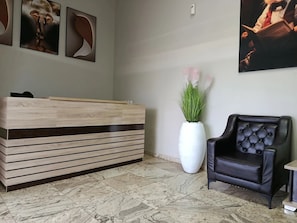 Un mostrador de recepción, un sillón de cuero negro, un jarrón grande con plantas verdes y tres cuadros en la pared.