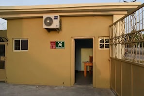 A building with a beige exterior, an air conditioning unit mounted on the wall, a door with a green sign indicating an assembly point, and a barred gate on the right. - Mata Garden Hotel (Isalu)