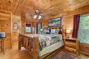 A wooden cabin bedroom with a bed, a ceiling fan, a nightstand, a lamp, a window with curtains, and a painting on the wall. - All Time High (Sevierville)