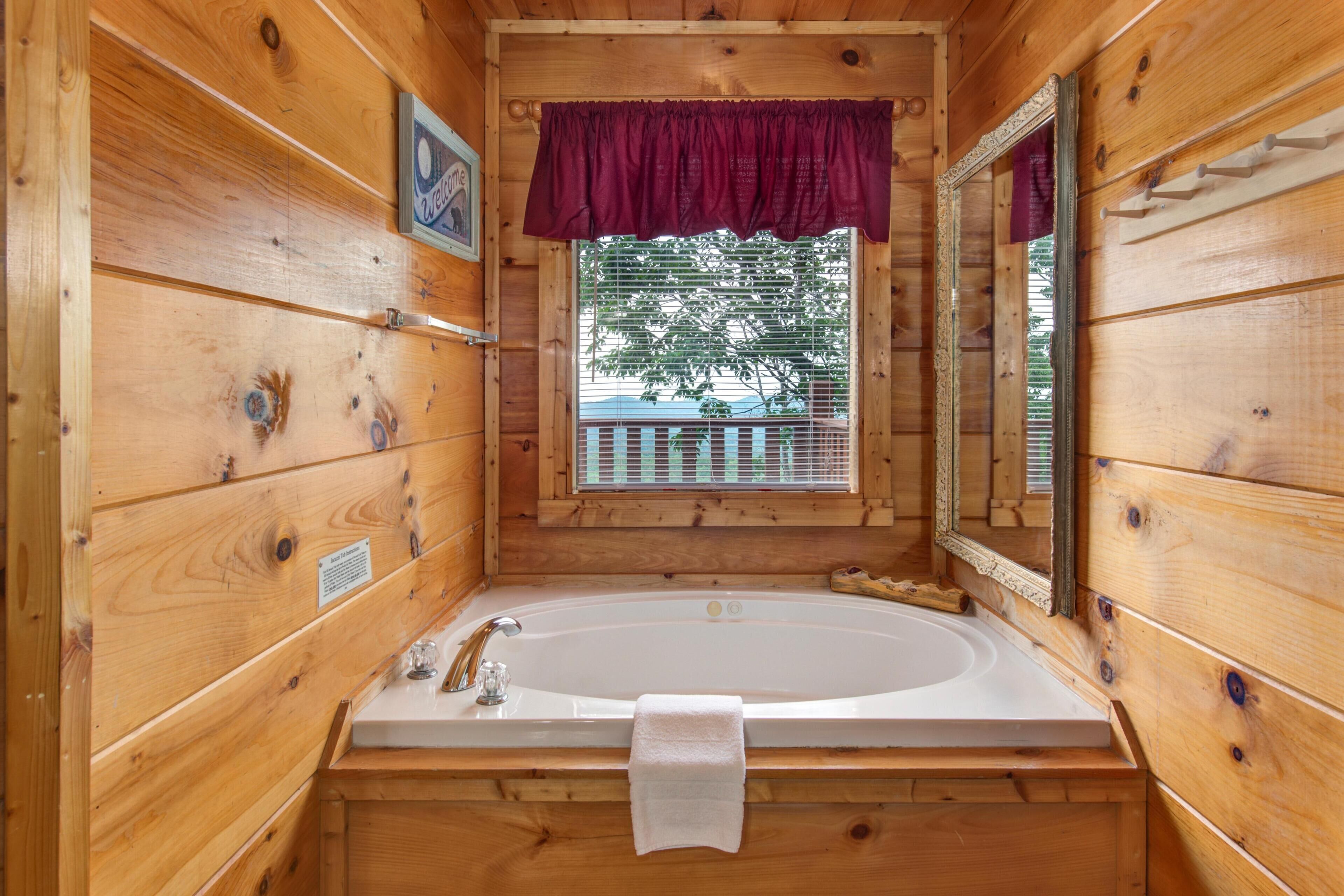 A wooden cabin bathroom with a large bathtub, a window with a view of a tree, and a mirror.