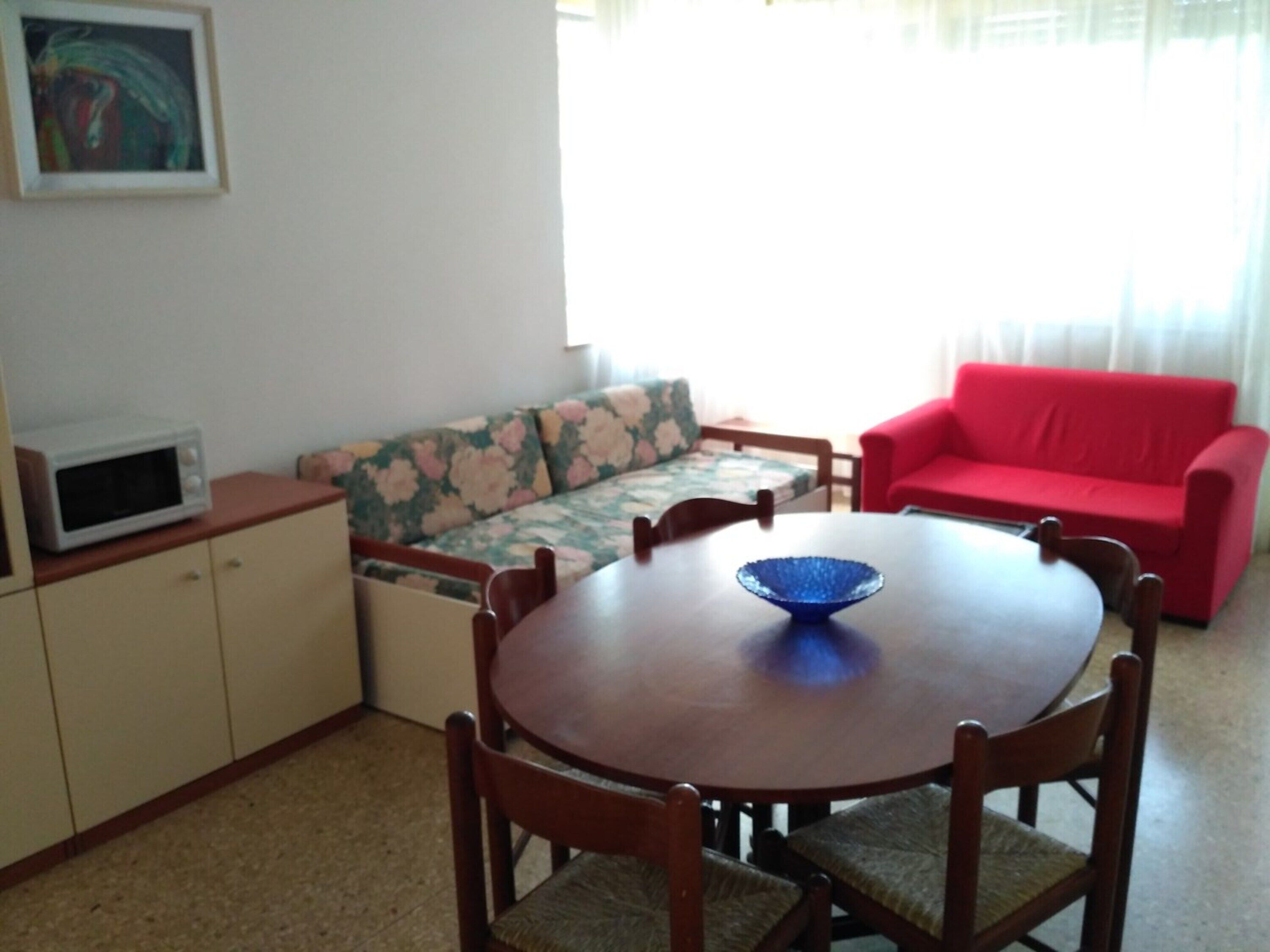 Zona pranzo con tavolo rotondo, sedie, un divano rosso, un forno a microonde e un divano con motivo floreale.