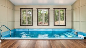 En swimmingpool med træterrasse, tre vinduer med trærammer og udsigt til et skovområde.