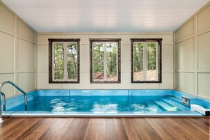 A swimming pool with wooden deck flooring, three windows with wooden frames, and a view of a forest.