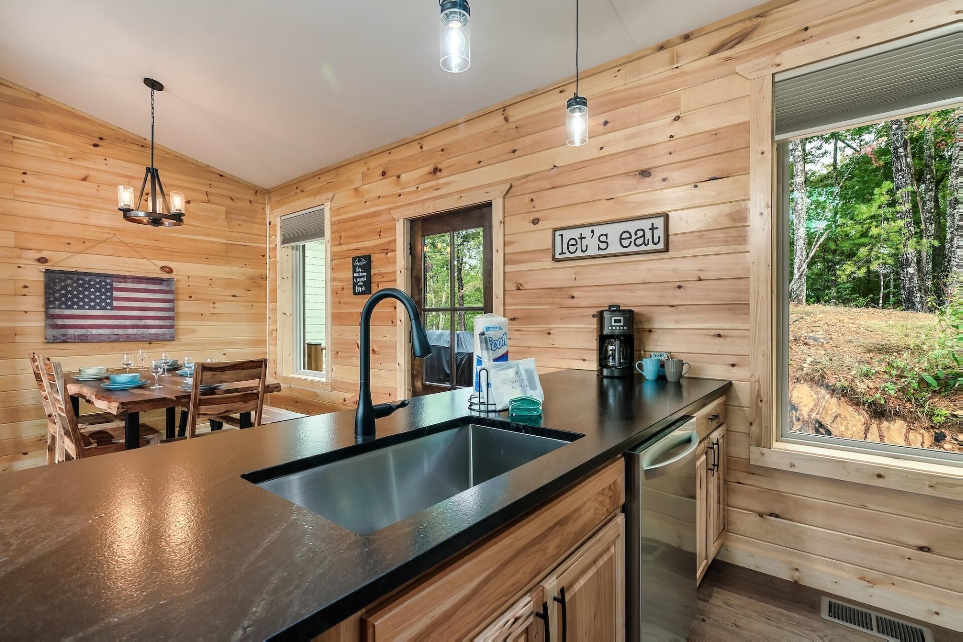 Interior de una cabaña de madera con zona de cocina, mesa de comedor y vistas al exterior.