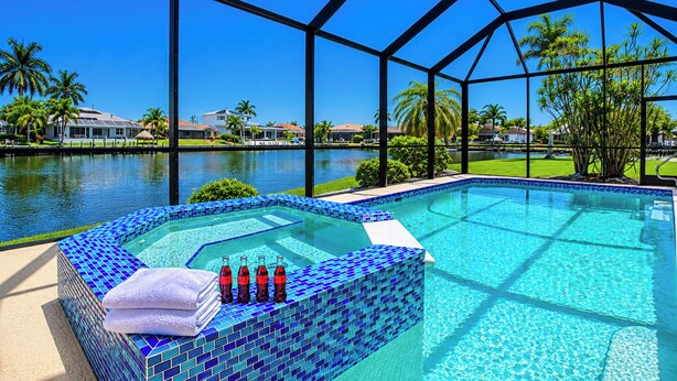Une piscine avec un bord carrelé, un jacuzzi et une vue sur un lac bordé de maisons et de palmiers.