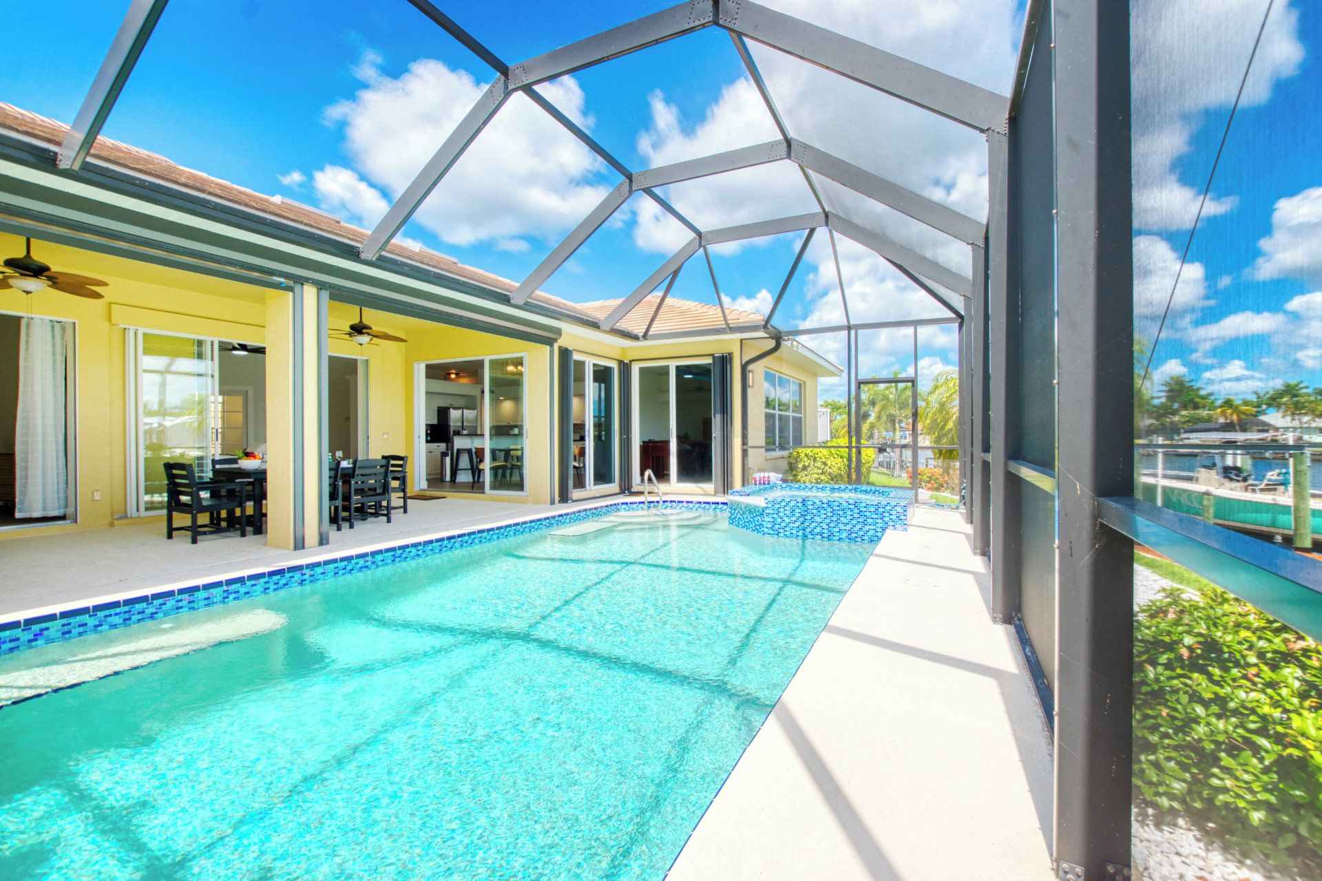 Une piscine dotée d’une structure en verre, un espace patio aménagé avec du mobilier d’extérieur et une vue sur un plan d’eau.