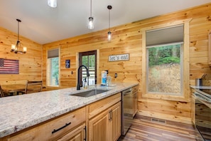 Interior de una cabaña de madera con zona de cocina, que incluye encimera de granito, fregadero de acero inoxidable y un ventanal con vistas a un área boscosa.