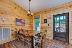 A wooden cabin interior with a dining table, chairs, a chandelier, an American flag, a door, and a window.