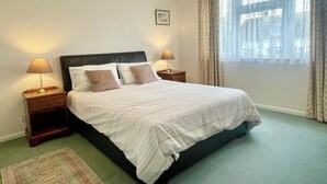 Uma cama bem arrumada com lençóis brancos, duas mesas de cabeceira com luminárias, um quadro na parede e uma janela grande com cortinas.