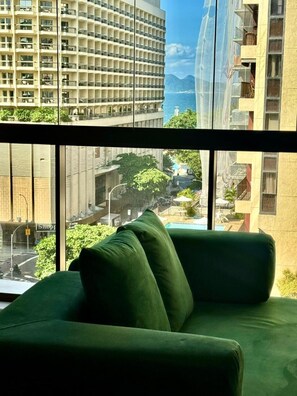 Blick aus einem Fenster auf eine Straße und Gebäude im Hintergrund, mit einem grünen Sofa im Vordergrund.