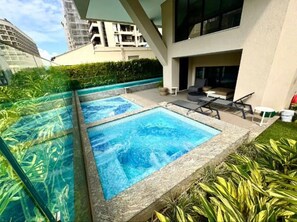 Ein modernes Außenpool-Areal mit zwei rechteckigen Pools, umgeben von viel Grün und an ein Gebäude mit Balkonen angrenzend.