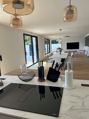 A modern kitchen with a built-in induction cooktop, a dining area with a long table and chairs, and a large window offering a view of the sea. - Villa Zoliette (Batz-sur-Mer)