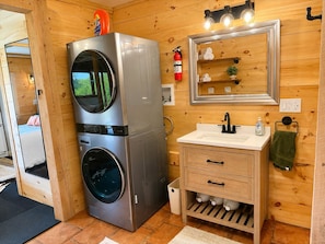 A compact bathroom with a washing machine, a sink, a mirror, and a small shelf with toiletries.