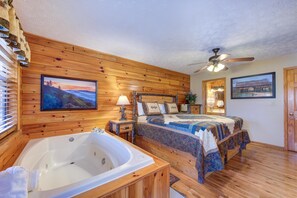 Quarto com painéis de madeira, uma cama grande, banheiro separado com banheira e um ventilador de teto.