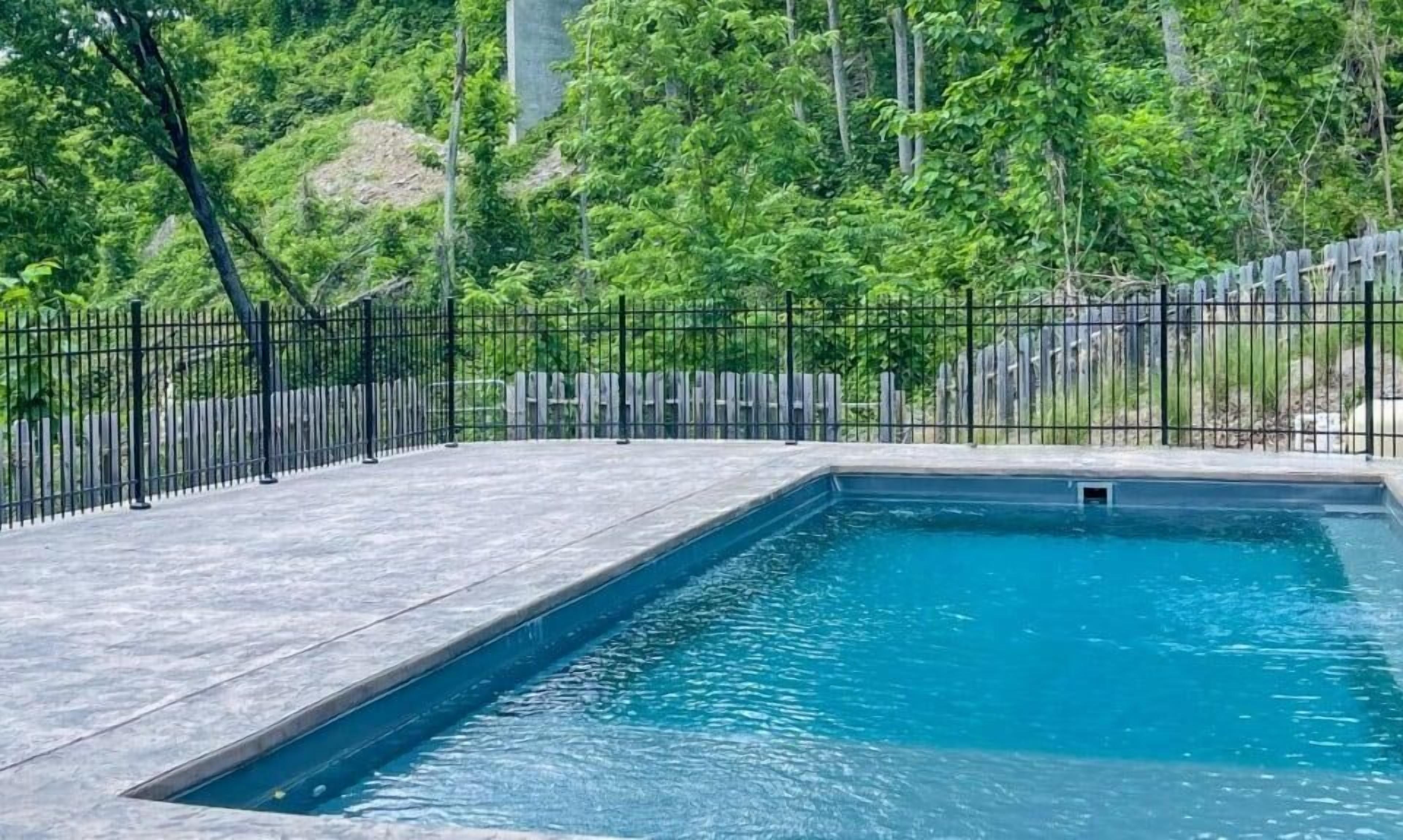 A rectangular swimming pool with a black metal fence, surrounded by a paved deck and lush greenery.