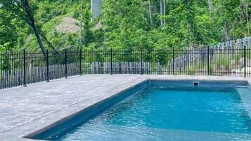 A rectangular swimming pool with a black metal fence, surrounded by a paved deck and lush greenery.