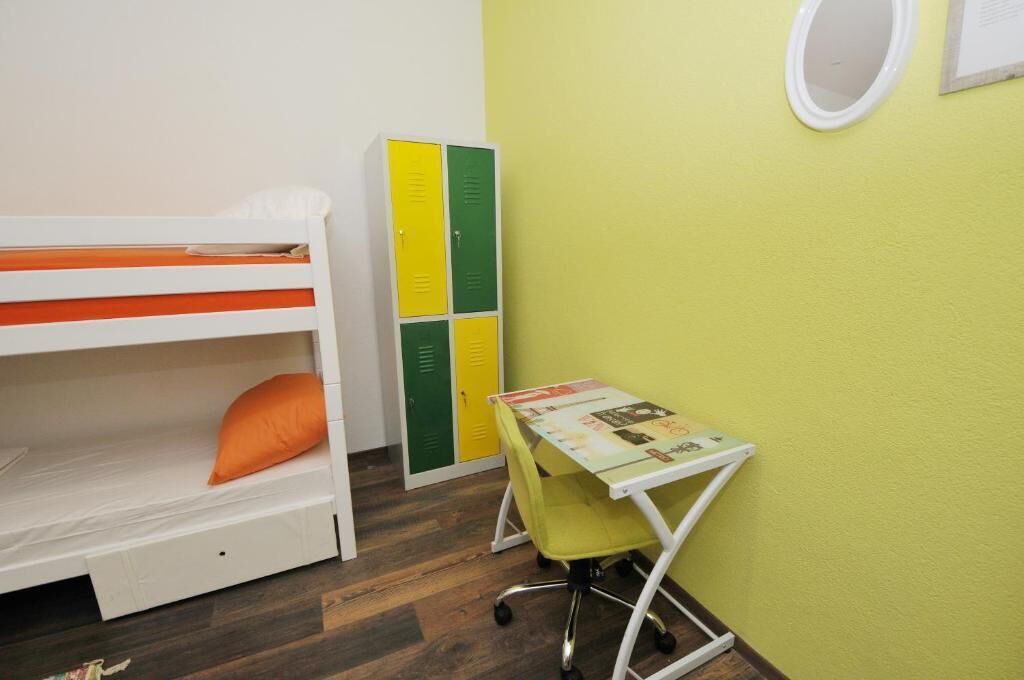 Una cama individual en literas con una almohada naranja, un escritorio pequeño con una silla y una serie de casilleros en verde y amarillo.