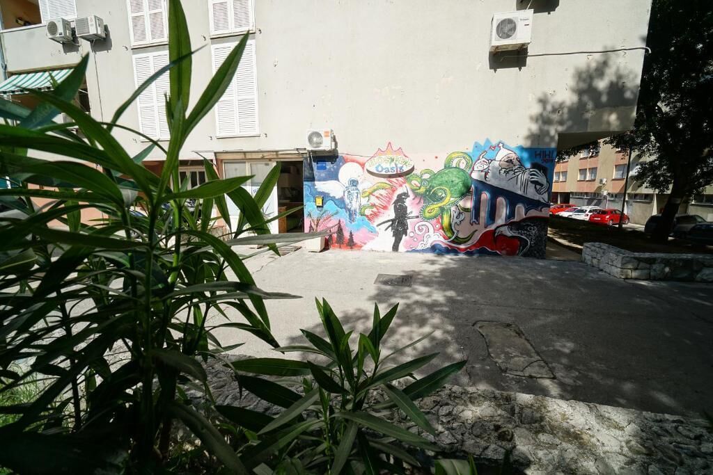 Un edificio con un mural colorido en la pared, un área pavimentada y algo de vegetación al frente.