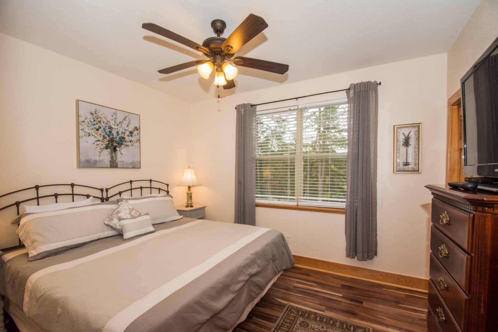 A bedroom with a bed, a ceiling fan, a window with curtains, a nightstand with a lamp, and a dresser.