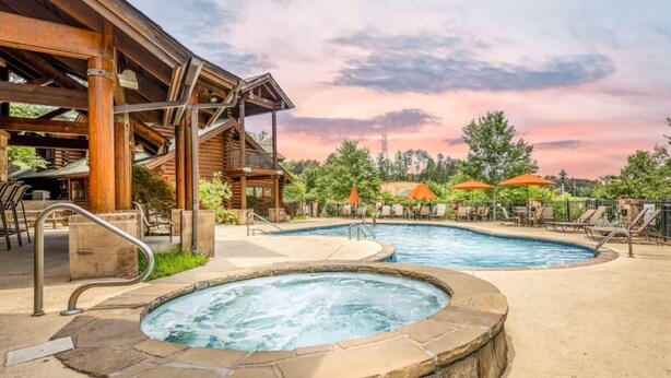 Una cabaña de madera con jacuzzi y piscina, rodeada de mobiliario exterior y sombrillas.