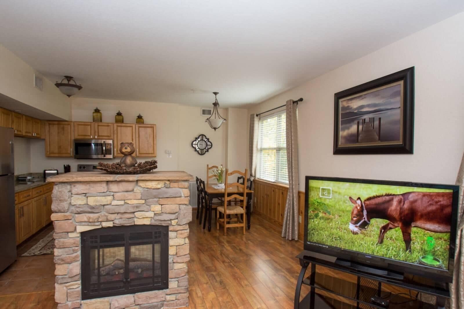 Una sala de estar con chimenea de piedra, mesa de comedor de madera y un televisor mostrando un caballo.