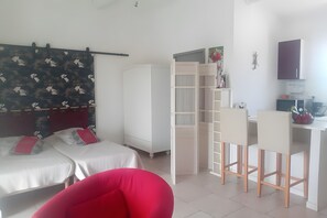 A room with two beds, a red chair, a white desk with two chairs, a wardrobe, and a small kitchenette.