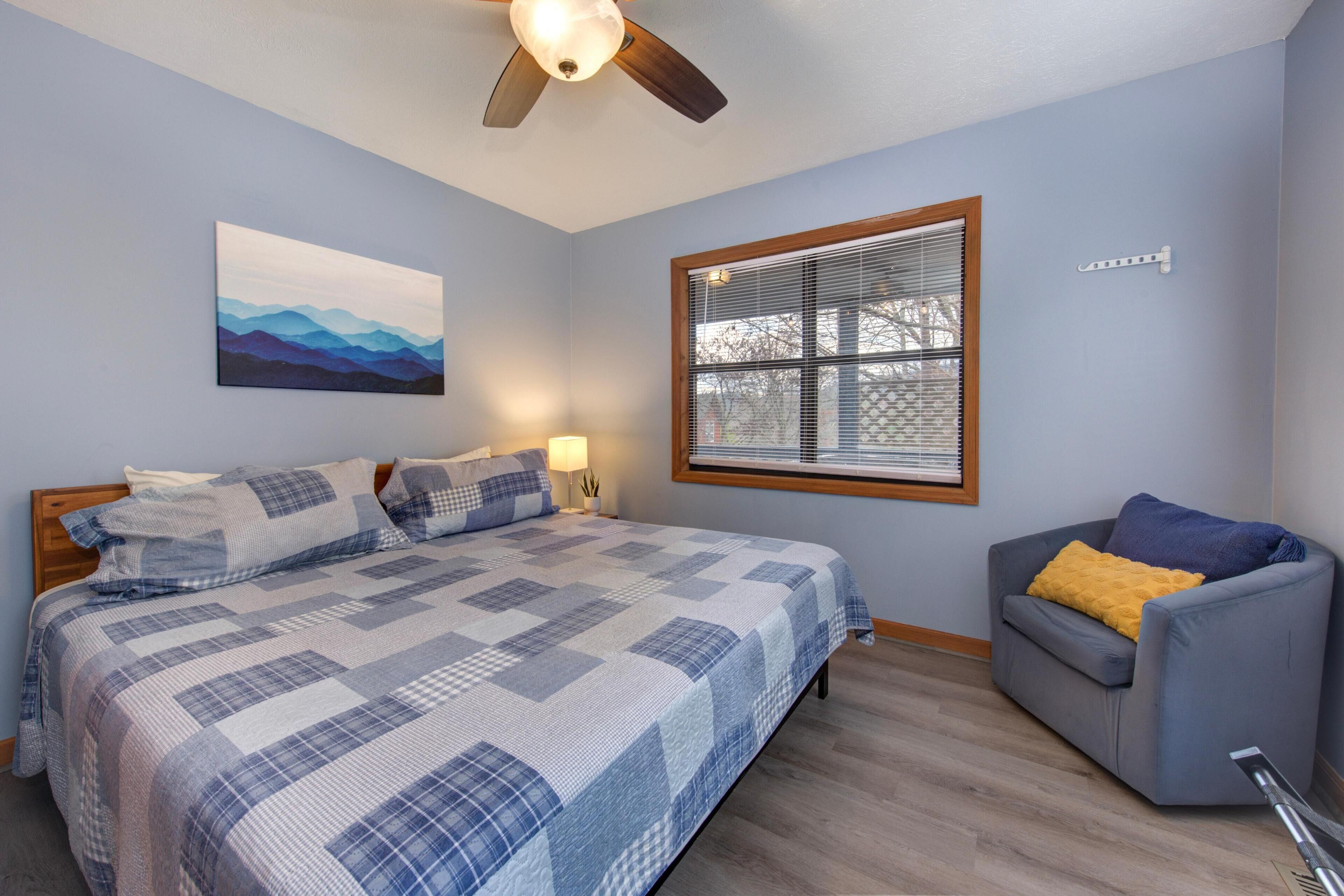 A bedroom with a bed, a window with blinds, a ceiling fan, and a painting on the wall.