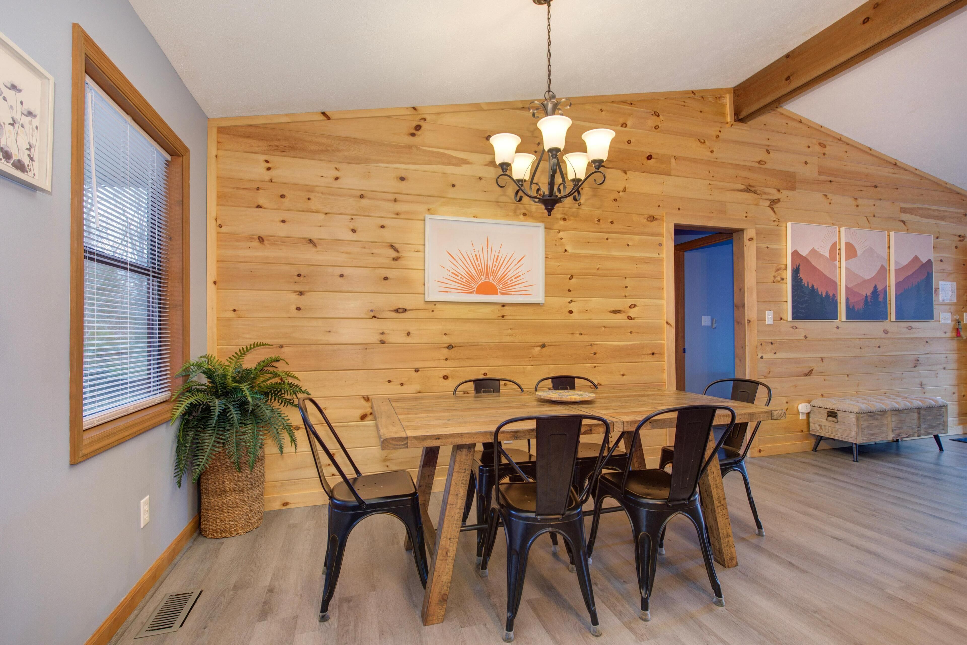 A wooden interior with a dining table, chairs, a chandelier, and artwork on the walls.