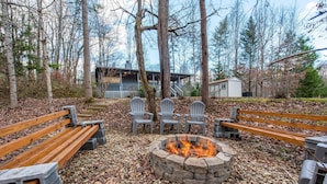 A fire pit surrounded by wooden benches and Adirondack chairs in a forested area with a house in the background. - Coyote Crossing (Sevierville)