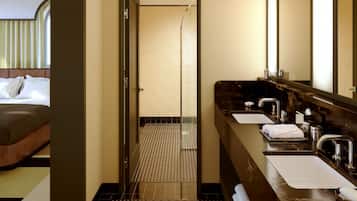 A hotel bathroom with a shower, two sinks, and a bed visible through an open door.