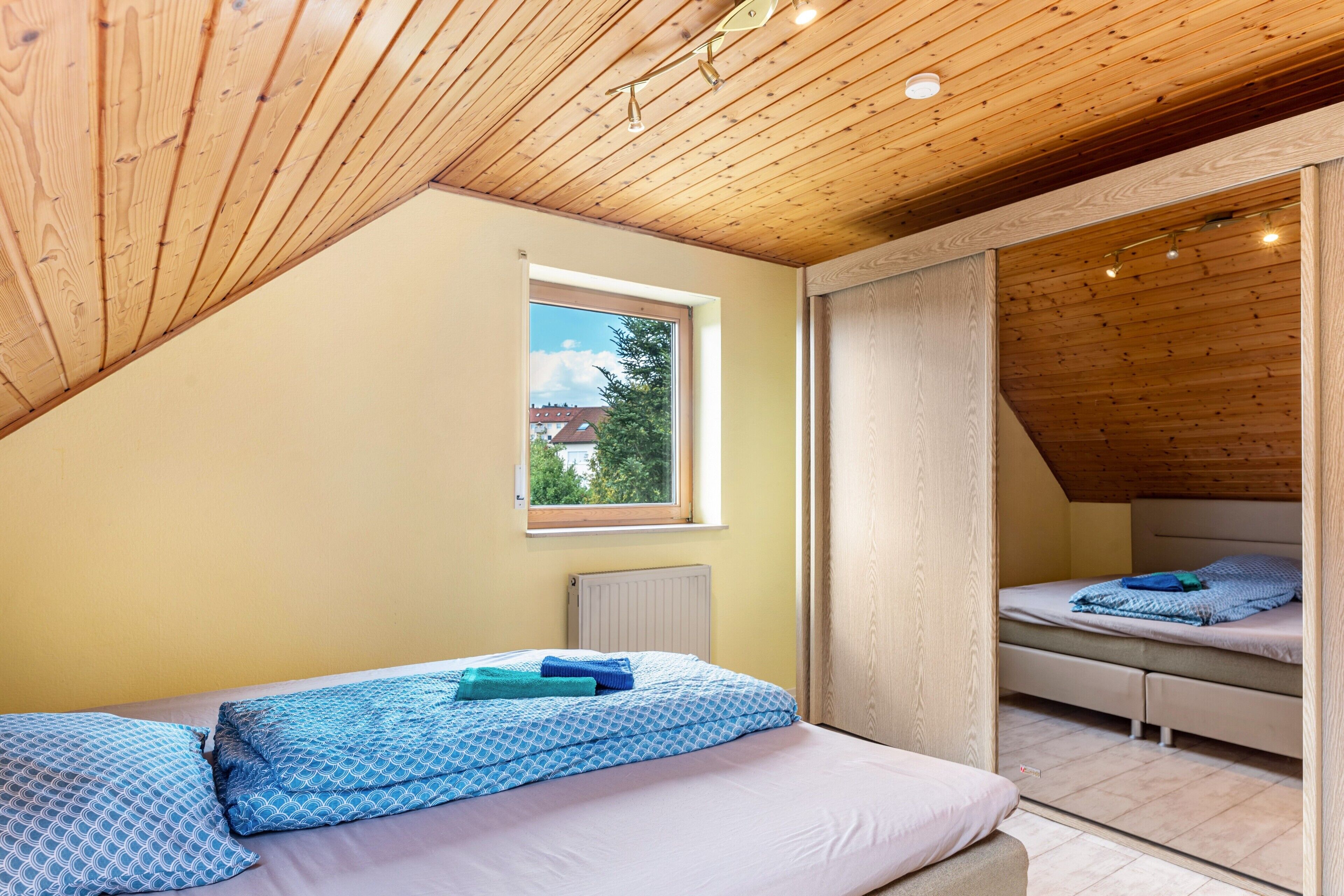 Een slaapkamer met een schuine kap, een raam met uitzicht en twee bedden met blauw beddengoed.