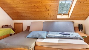 Een slaapkamer met schuine wand, een raam met uitzicht op bomen, een bed met kussens en een nachtkastje met een lamp.