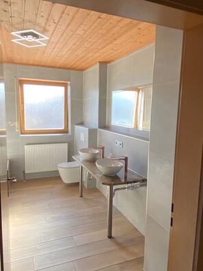 A modern bathroom with a wooden ceiling, a large window, a white toilet, and two vessel sinks.