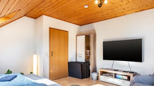 Een moderne kamer met een houten plafond, een flatscreen televisie aan de wand, een bed met een blauw beddegoed met een patroon, en een zithoek met een grijze bank.