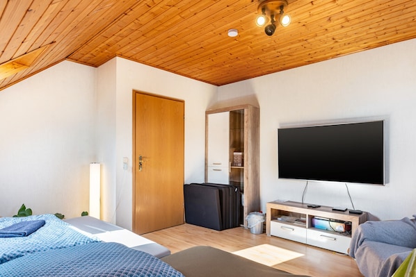 Una habitación moderna con techo de madera, televisor de pantalla plana montado en la pared, cama con colcha azul estampada y un pequeño rincón de descanso con un sofá gris.