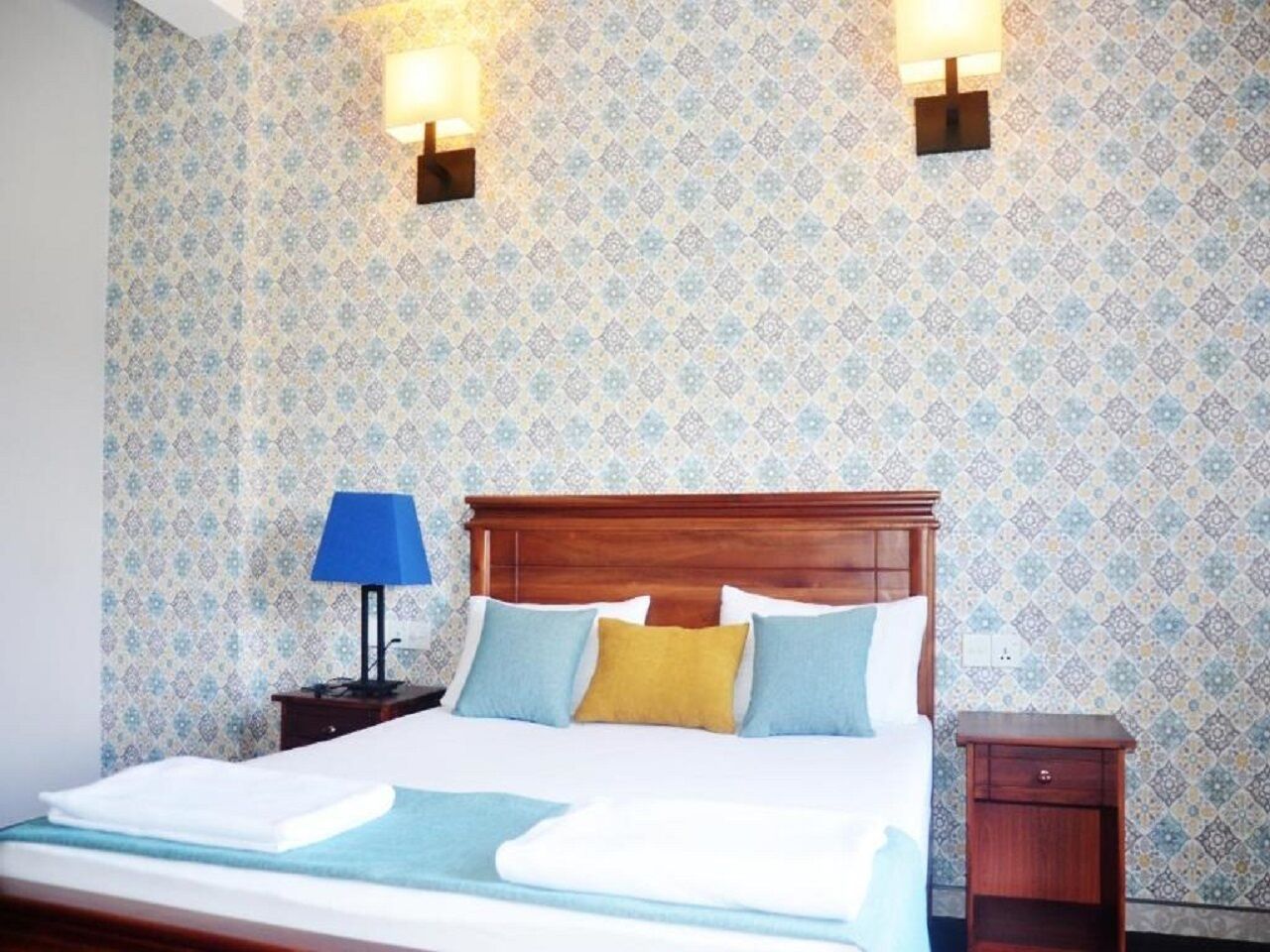 A neatly made bed with a wooden headboard, two bedside lamps, and a patterned wallpaper.