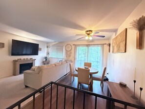 A living room with a fireplace, a sofa, a dining table with chairs, a ceiling fan, and a TV mounted on the wall.