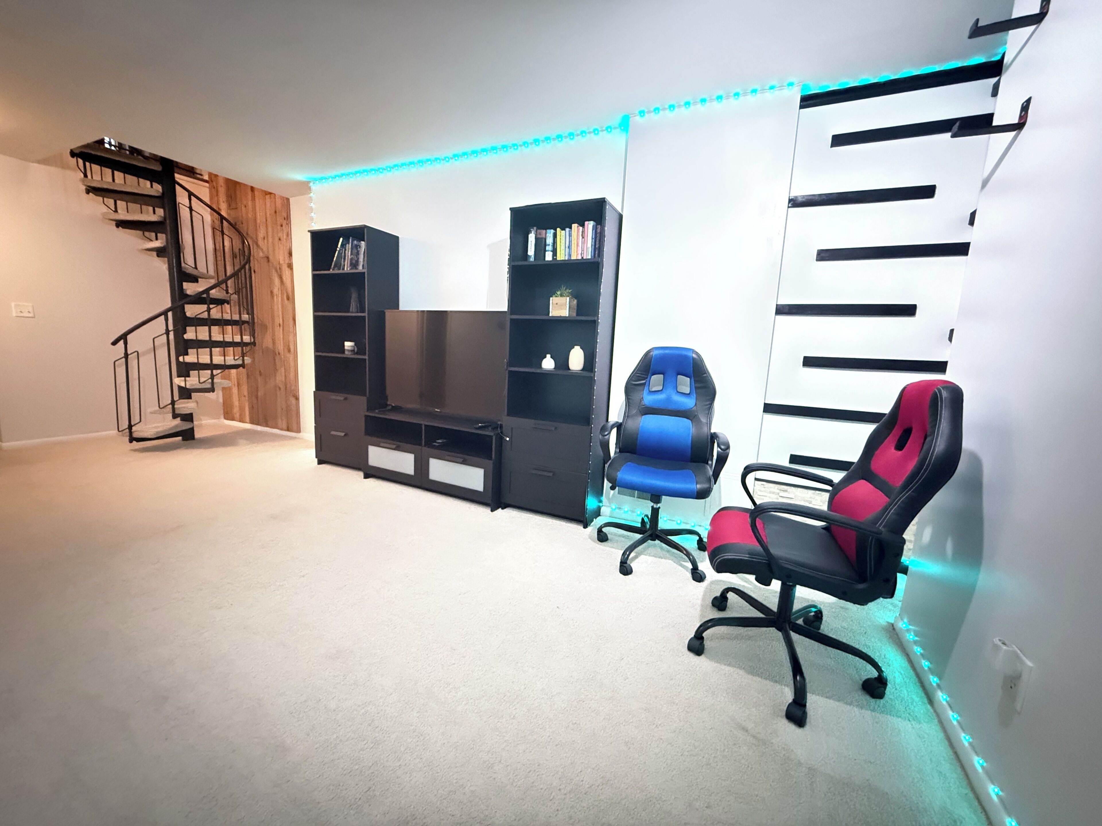 A room with a spiral staircase, black and white shelving units, a blue gaming chair, and a red gaming chair.