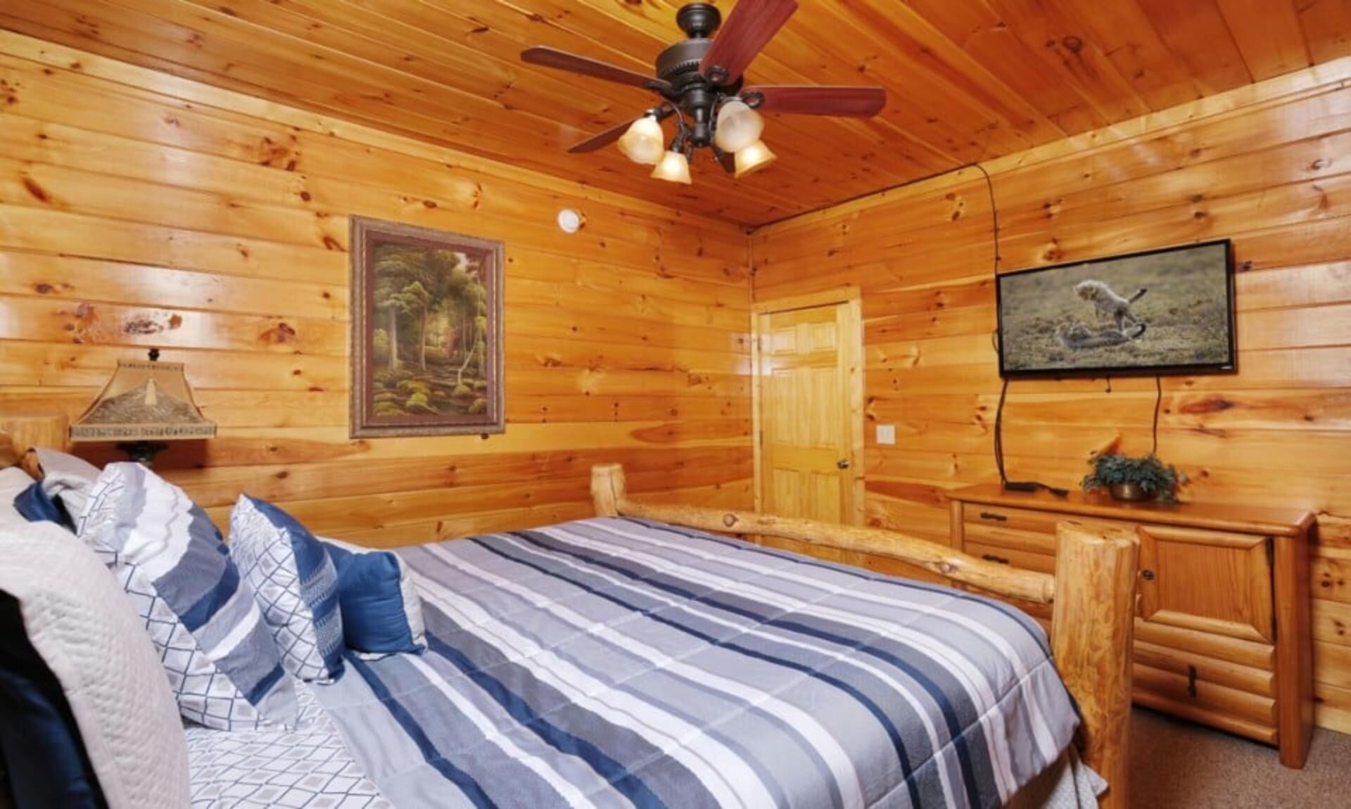 A wooden cabin bedroom with a bed, a ceiling fan, a mounted television, and a framed picture on the wall.