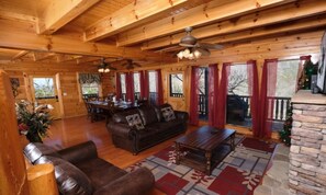 A log cabin interior with a dining area, a living room with a sofa and coffee table, and a stone fireplace. - Splash N Sunrise Theatre (Sevierville)