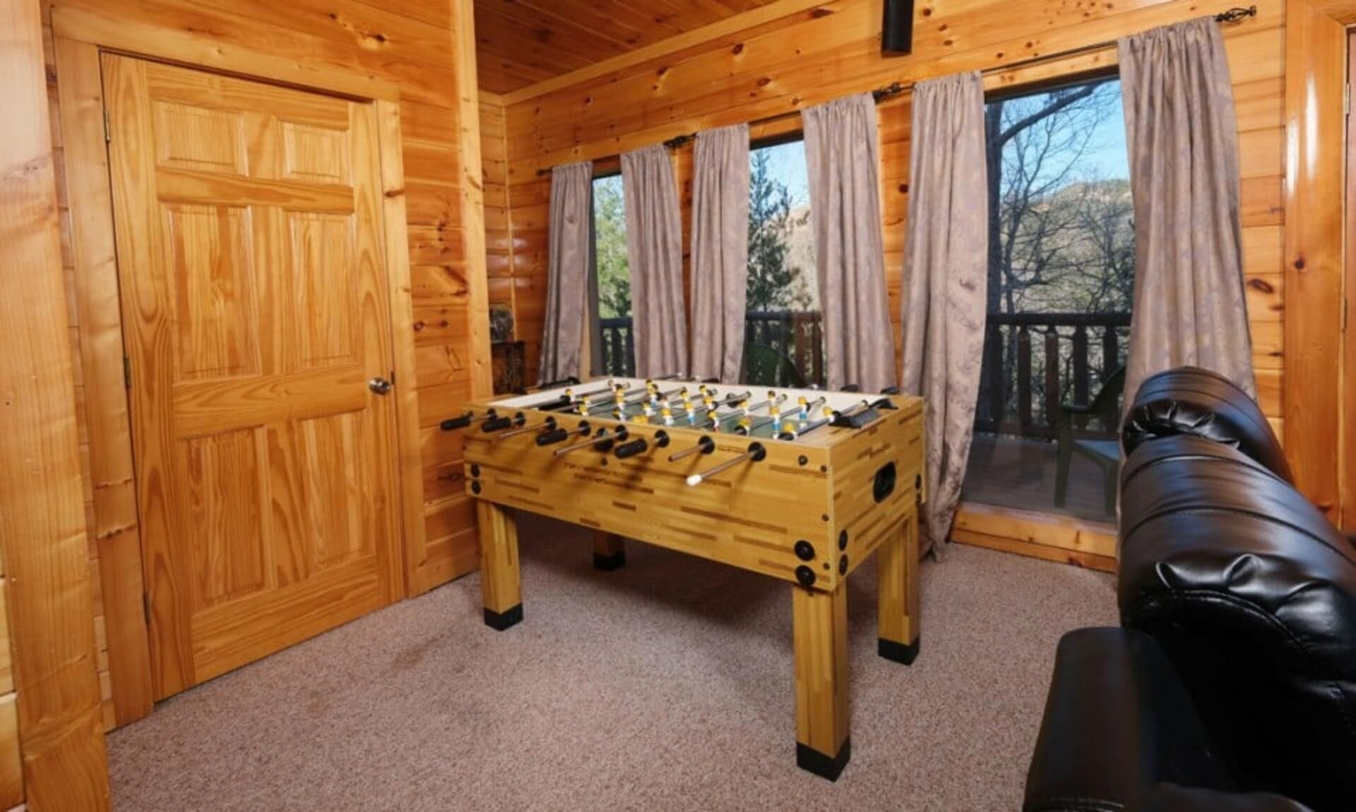 A wooden-floored room with a foosball table, a sofa, and a large window with curtains.