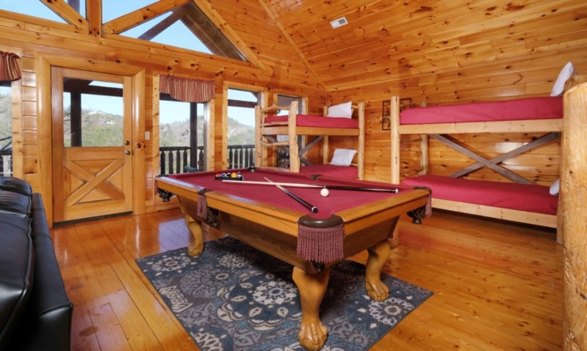 A wooden cabin with a pool table, bunk beds, and a view of the outdoors.