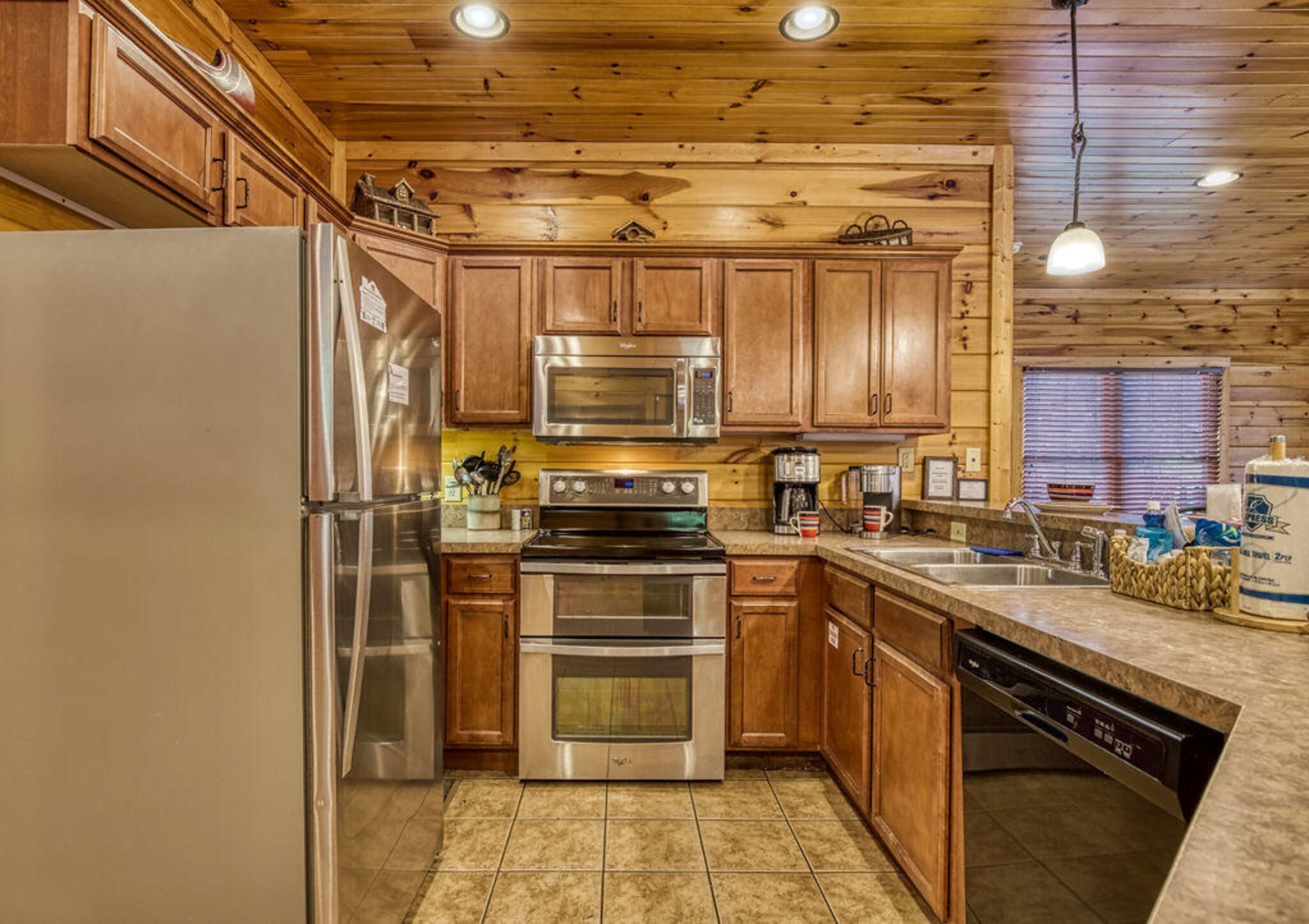Una cocina con armarios de madera, electrodomésticos de acero inoxidable, lavavajillas y una ventana con persianas.
