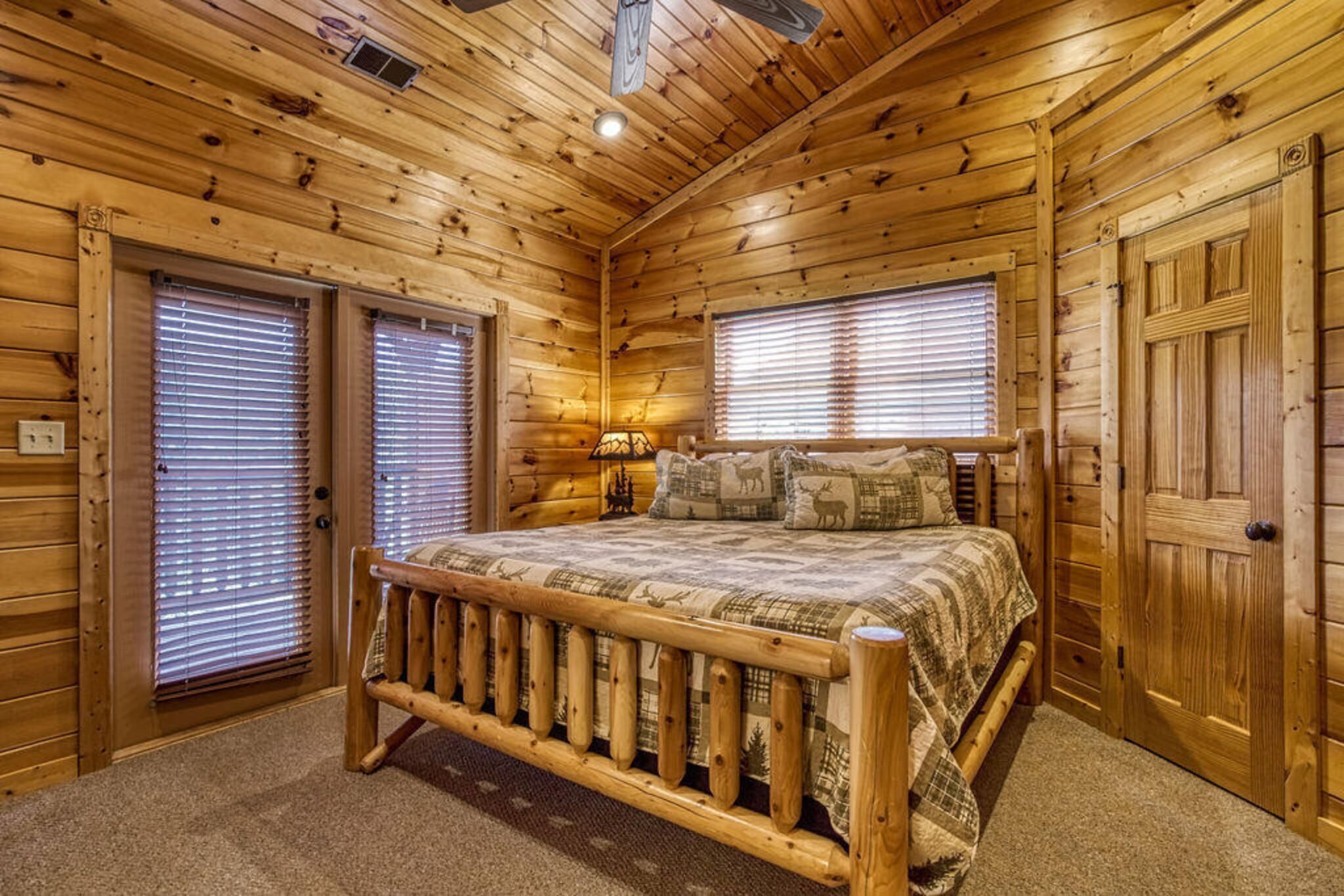 Un dormitorio tipo cabaña de madera con una cama de madera, dos ventanas con persianas, una puerta y un ventilador de techo.