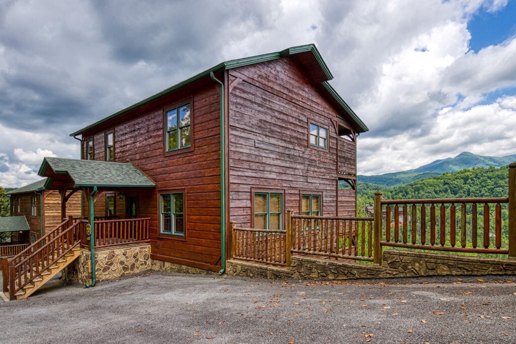 A two-story wooden cabin with a stone foundation, a wooden porch, and a stone wall.